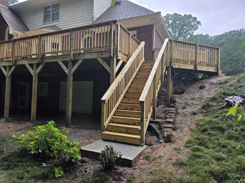 Wooden deck with stairs leading down to a concrete landing on a grassy hill; the house is behind the deck.