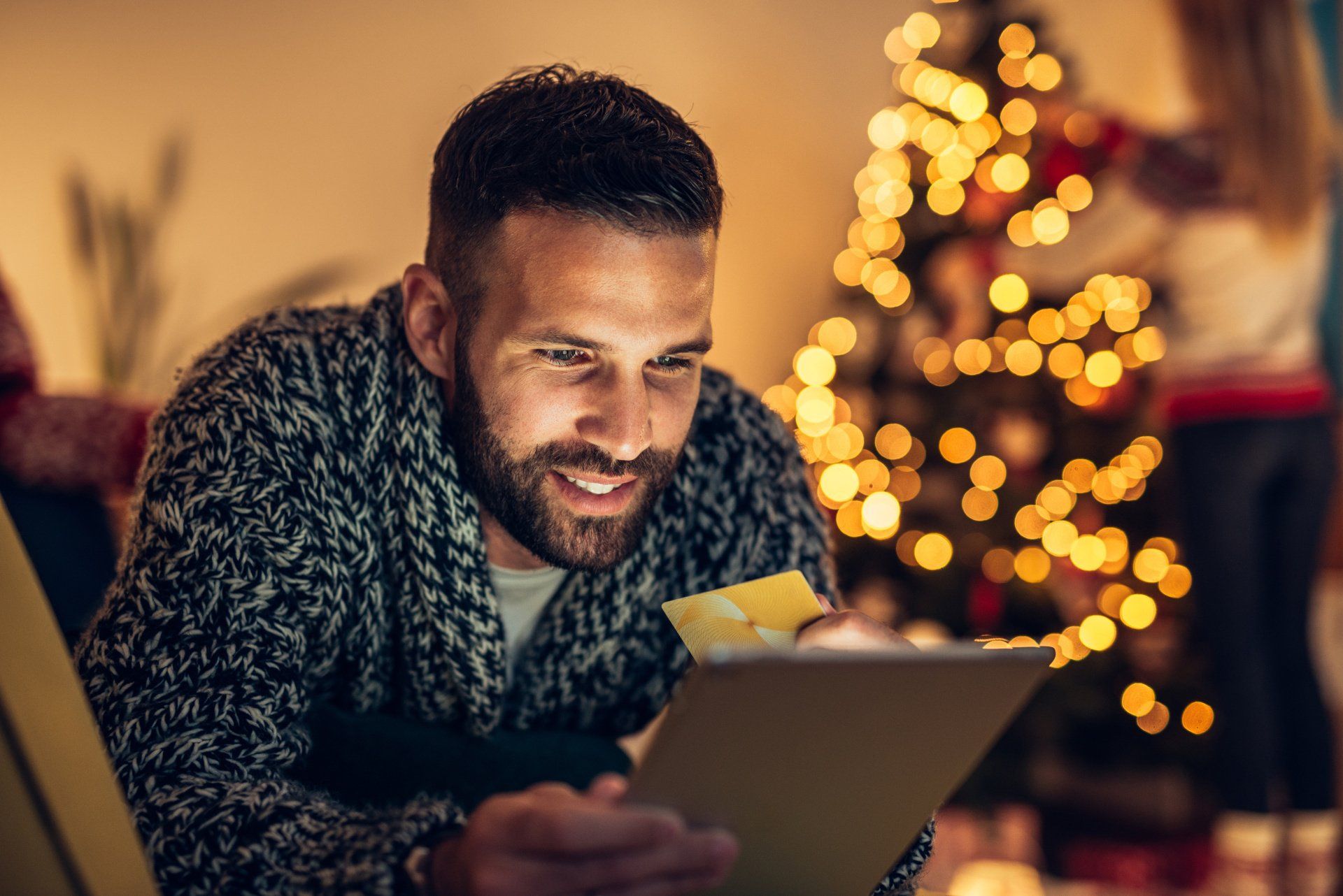 Image of a man holiday shopping online with a lit-up Christmas tree in a cozy and warm background