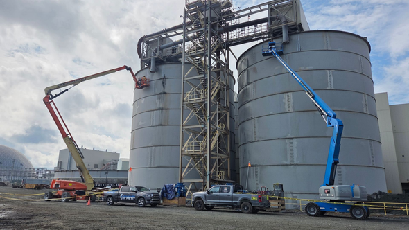 Two large gray industrial silos with construction equipment, under a cloudy sky.