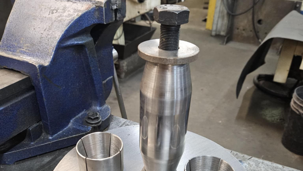 A metal cylinder being pressed with a bolt and nut assembly, held in a vise.