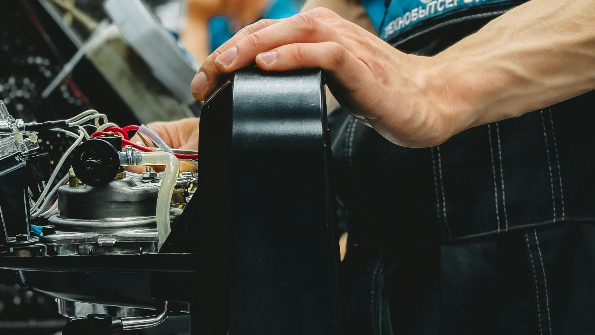 Person's hands assembling equipment, pressing against a black component. Worn apron visible.