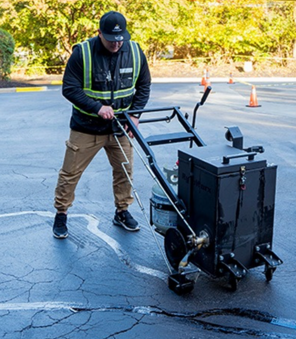Man in safety vest seals a parking lot crack with a black machine.
