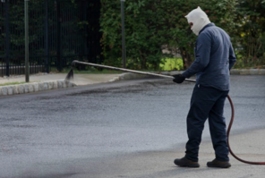 A person spraying a black substance on a paved surface, wearing protective clothing in an outdoor setting.