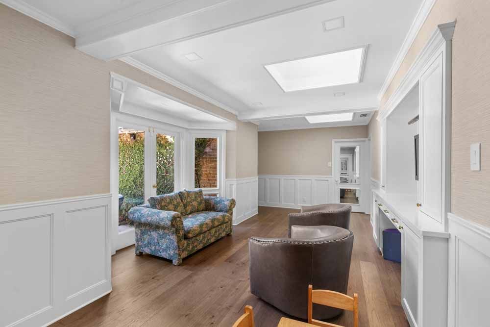 Spacious room with dark wood floors, white wainscoting, a couch, two chairs, and a window overlooking greenery.