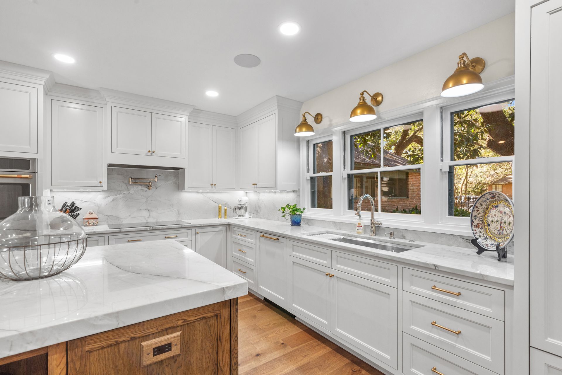 White kitchen with marble countertops, wooden island, and brass light fixtures.