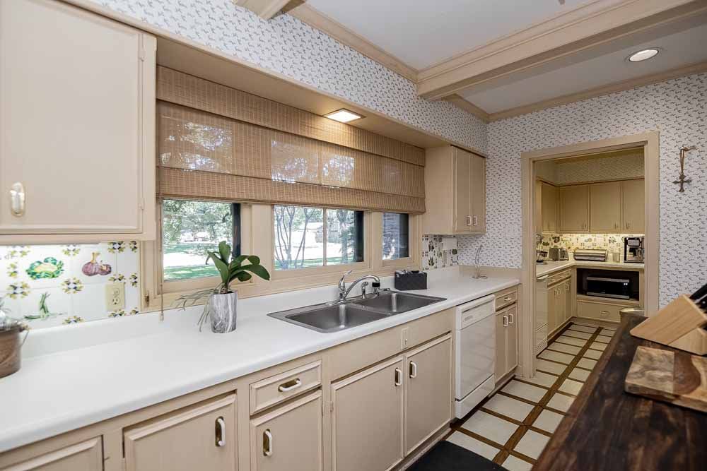 Beige kitchen with countertop, cabinets, window with shade, and a door to another room.
