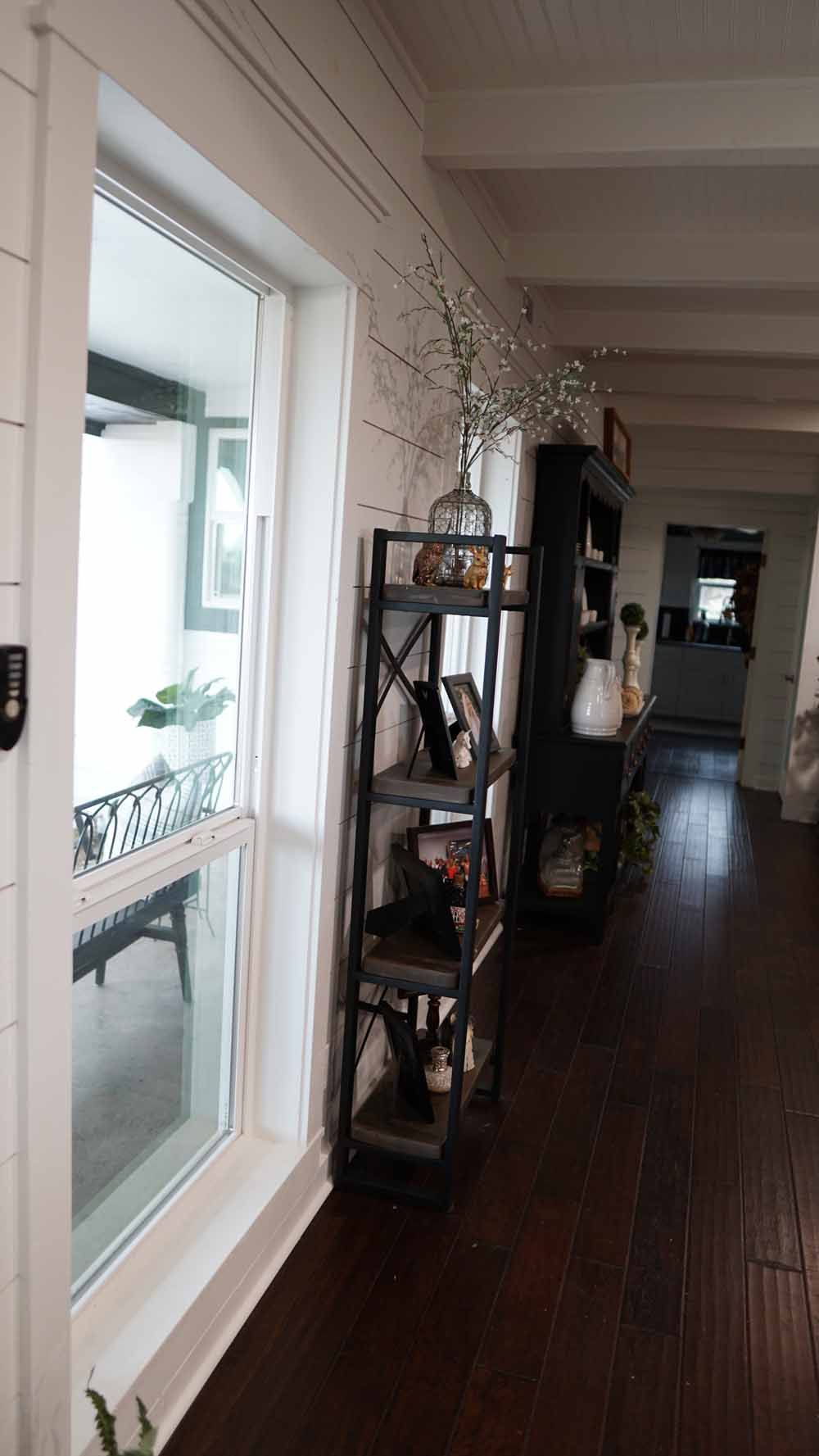 Dark wood floor, tall window, metal shelving unit with decor, white shiplap wall.