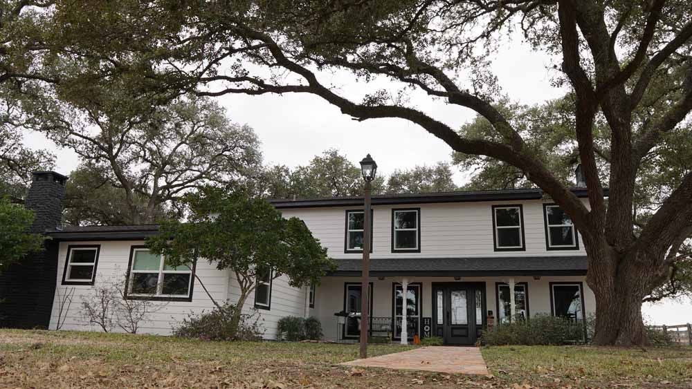 Two-story white house with black trim, a large tree in the front yard, and a lamppost on a cloudy day.