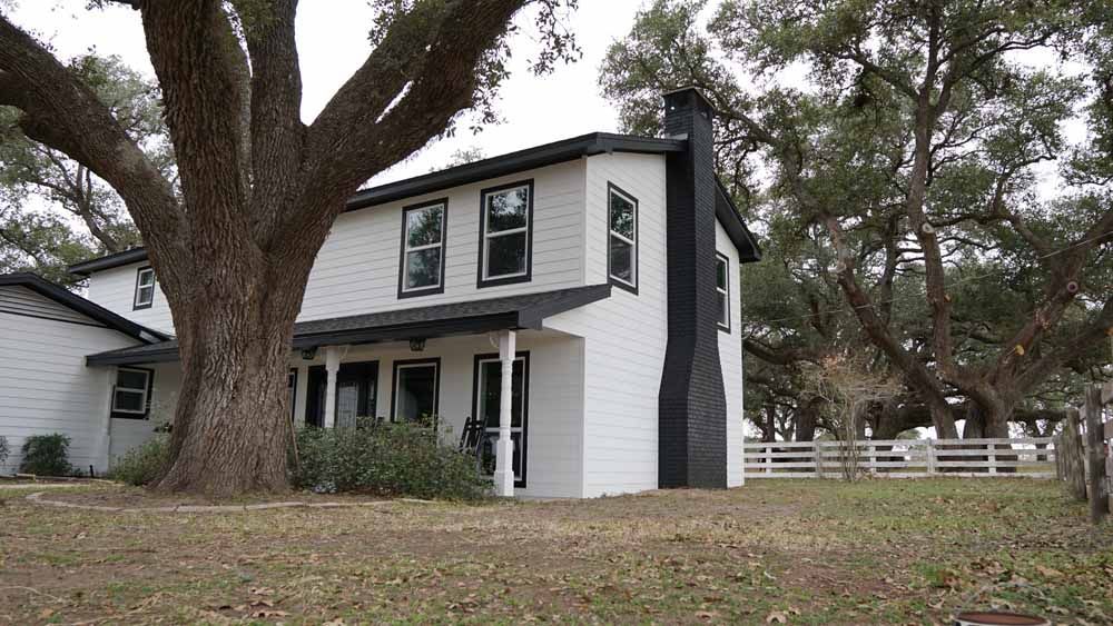 White two-story house with black trim, chimney, and porch. Large tree in front, grassy yard, and wooden fence.