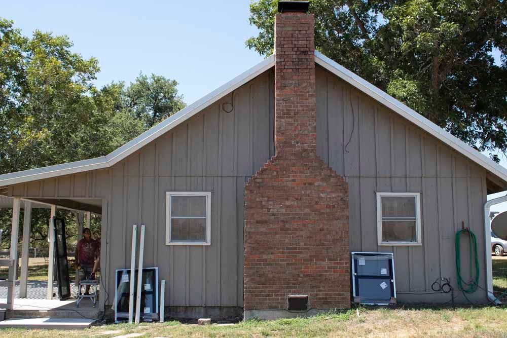 Tan house with a brick chimney and two windows. A person is under the porch, with blue panels leaning against the house.