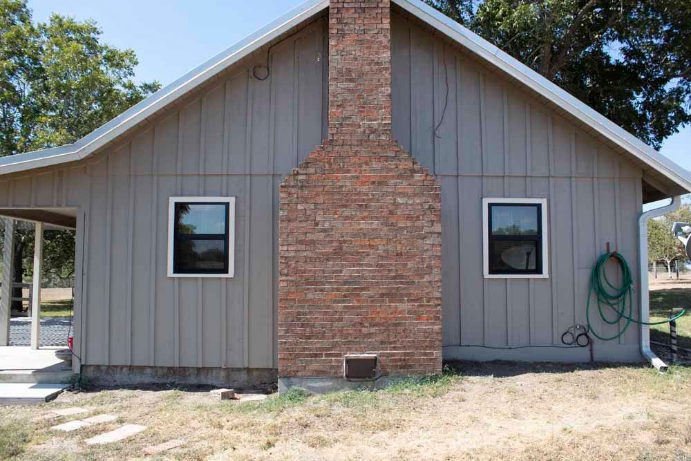Beige house with brick chimney, two windows, and a green garden hose.