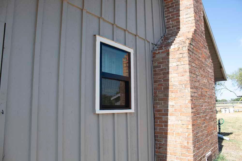 Side of a building with a window and brick chimney. Vertical gray siding, white window trim, and a clear sky.