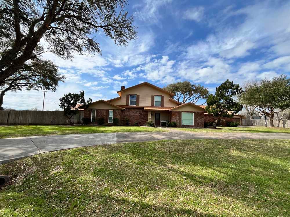 Two-story house with brick exterior and brown roof, set on a green lawn with trees and a cloudy blue sky.
