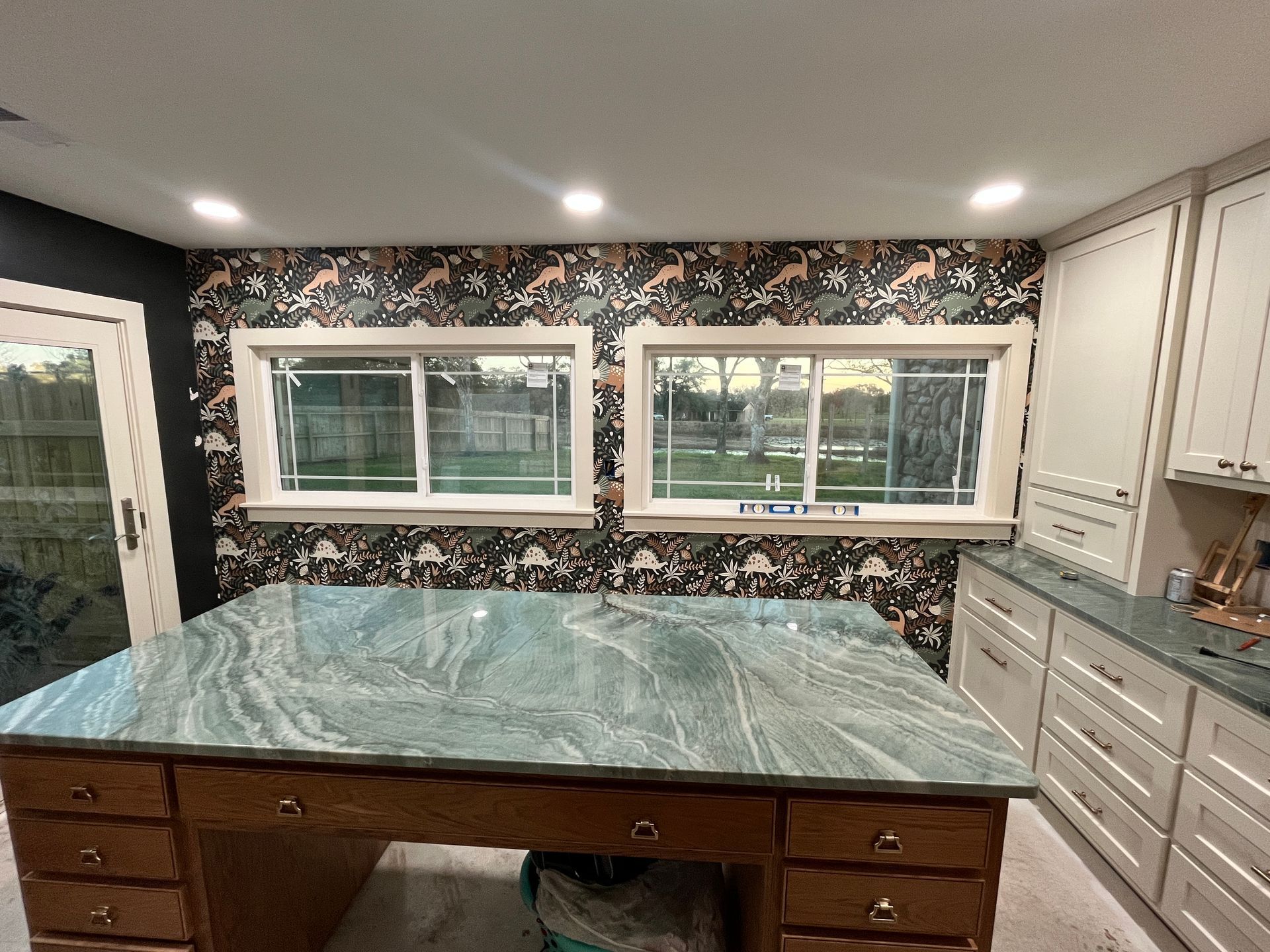 A room with a large desk and granite countertop, windows with decorative wallpaper, and cabinetry.