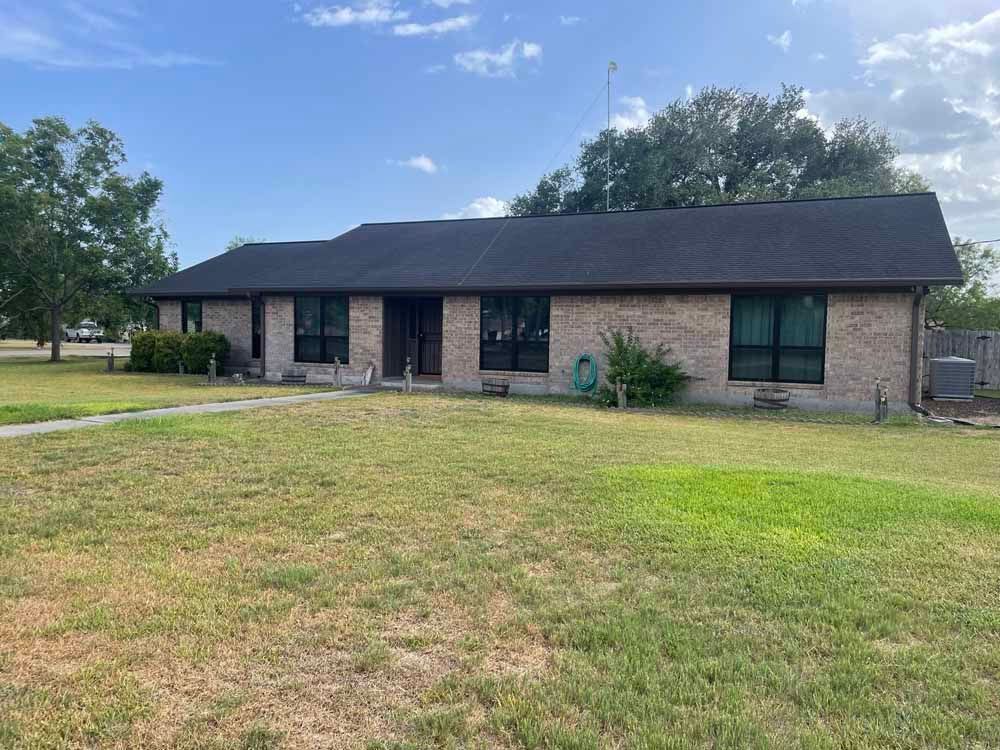 Brick ranch-style house with dark roof and windows, set on a grassy lawn under a partly cloudy sky.