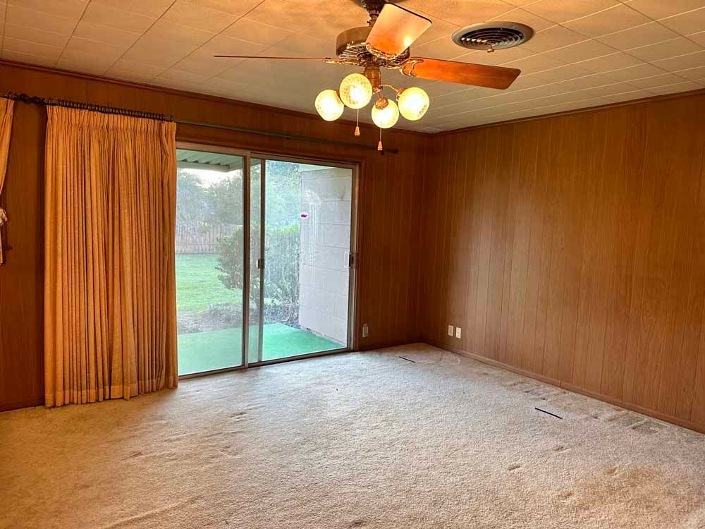 Empty room with tan carpet, wood paneling, sliding glass door, and ceiling fan.