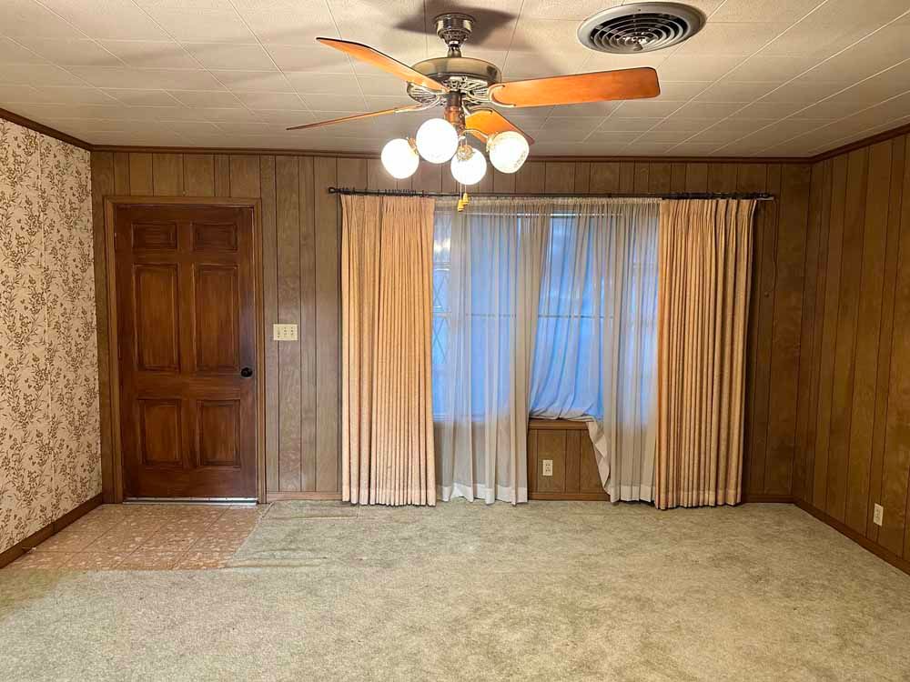 Empty living room with wood paneling, beige carpet, curtains, and a ceiling fan.