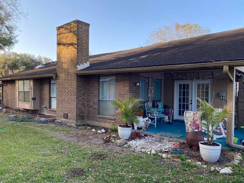 Brick home with chimney, covered patio, lawn, and potted plants.