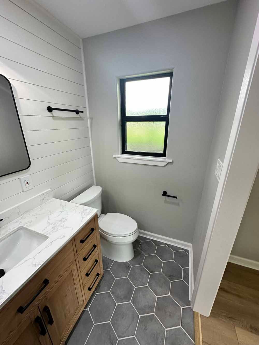 Bathroom with gray hexagonal tile floor, white toilet, wooden vanity, and a window.