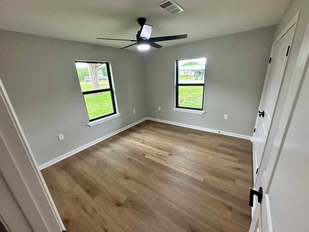Empty bedroom with light grey walls, wood-look floor, windows, and ceiling fan.