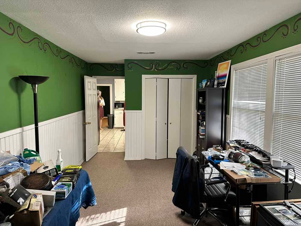 Room with green walls, white wainscoting, and a cluttered desk. A door leads to a kitchen.