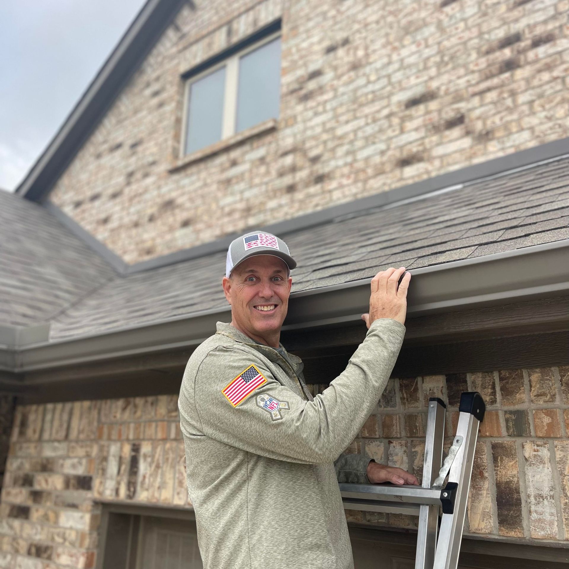 Man on a ladder cleaning a gutter on a brick house; smiling.