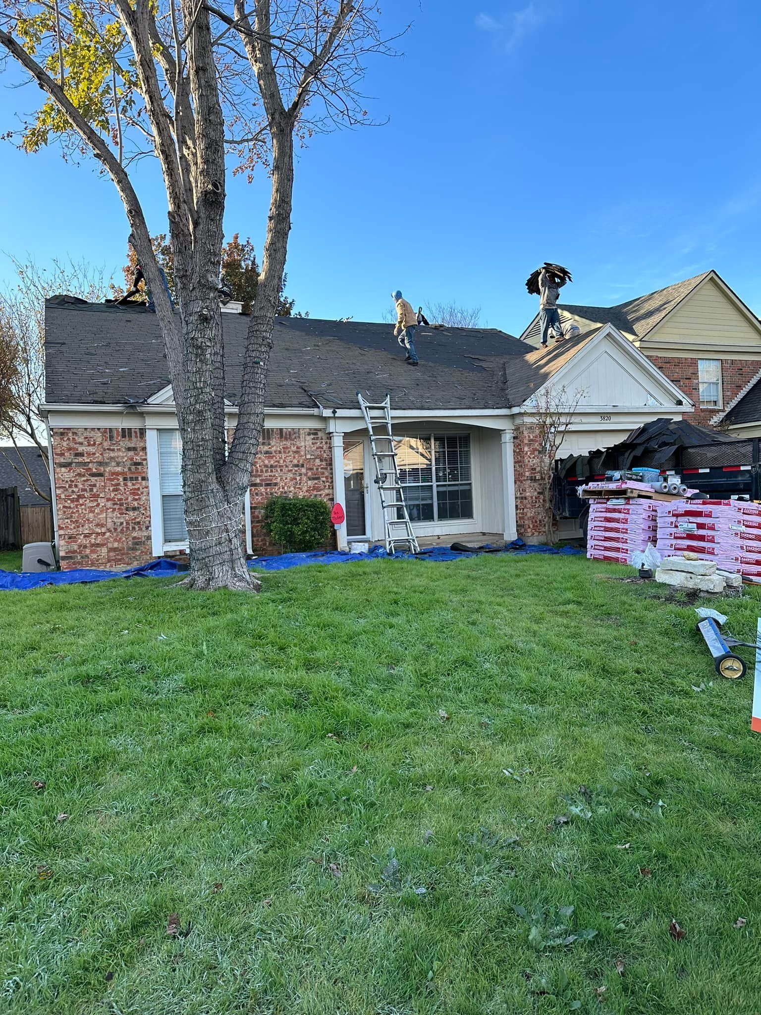 House with brick facade, roof under construction. Ladders, materials on lawn, tree in front. Blue sky.