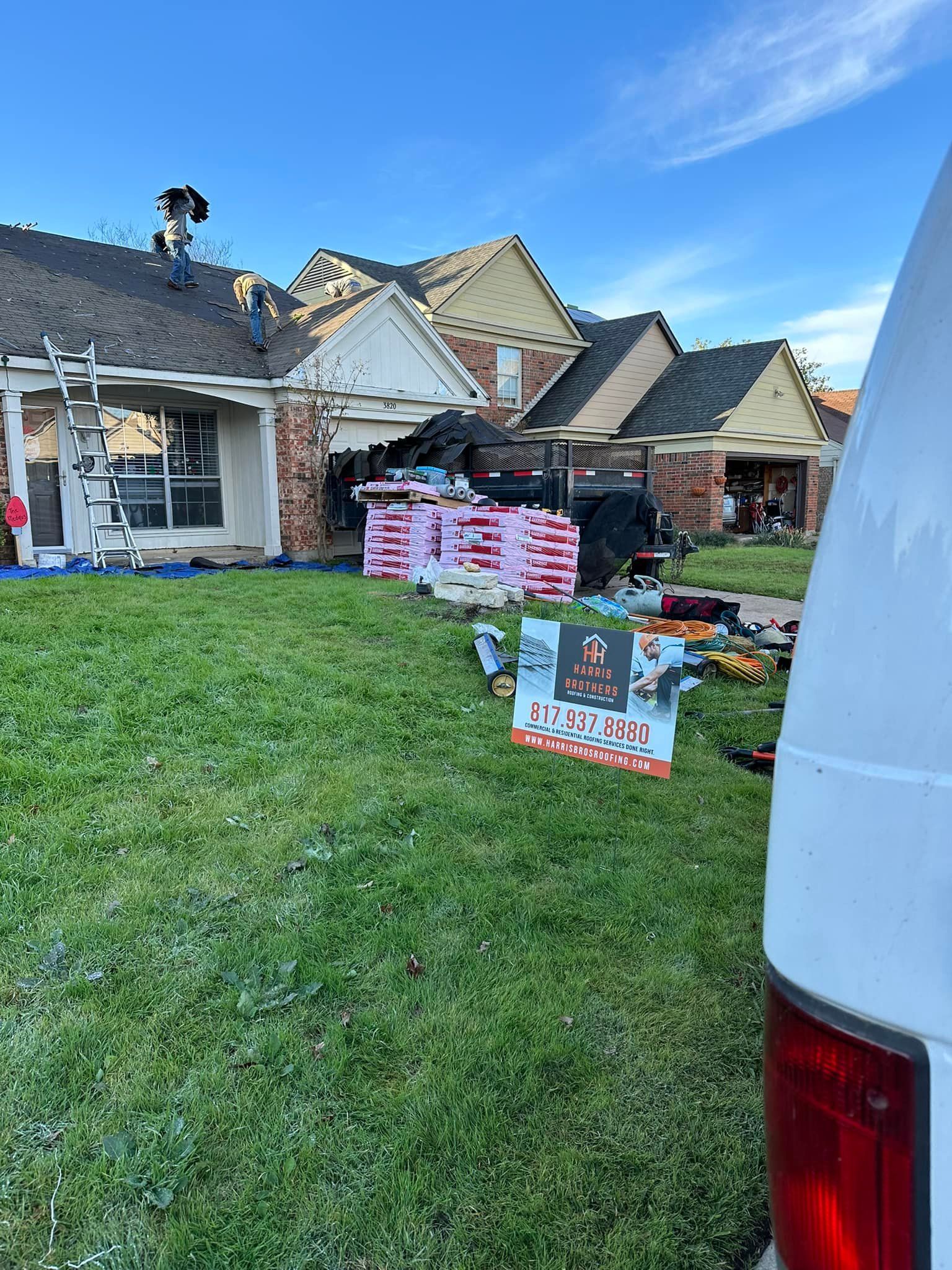 Roofing crew working on a house with a pile of shingles on the lawn under a blue sky.
