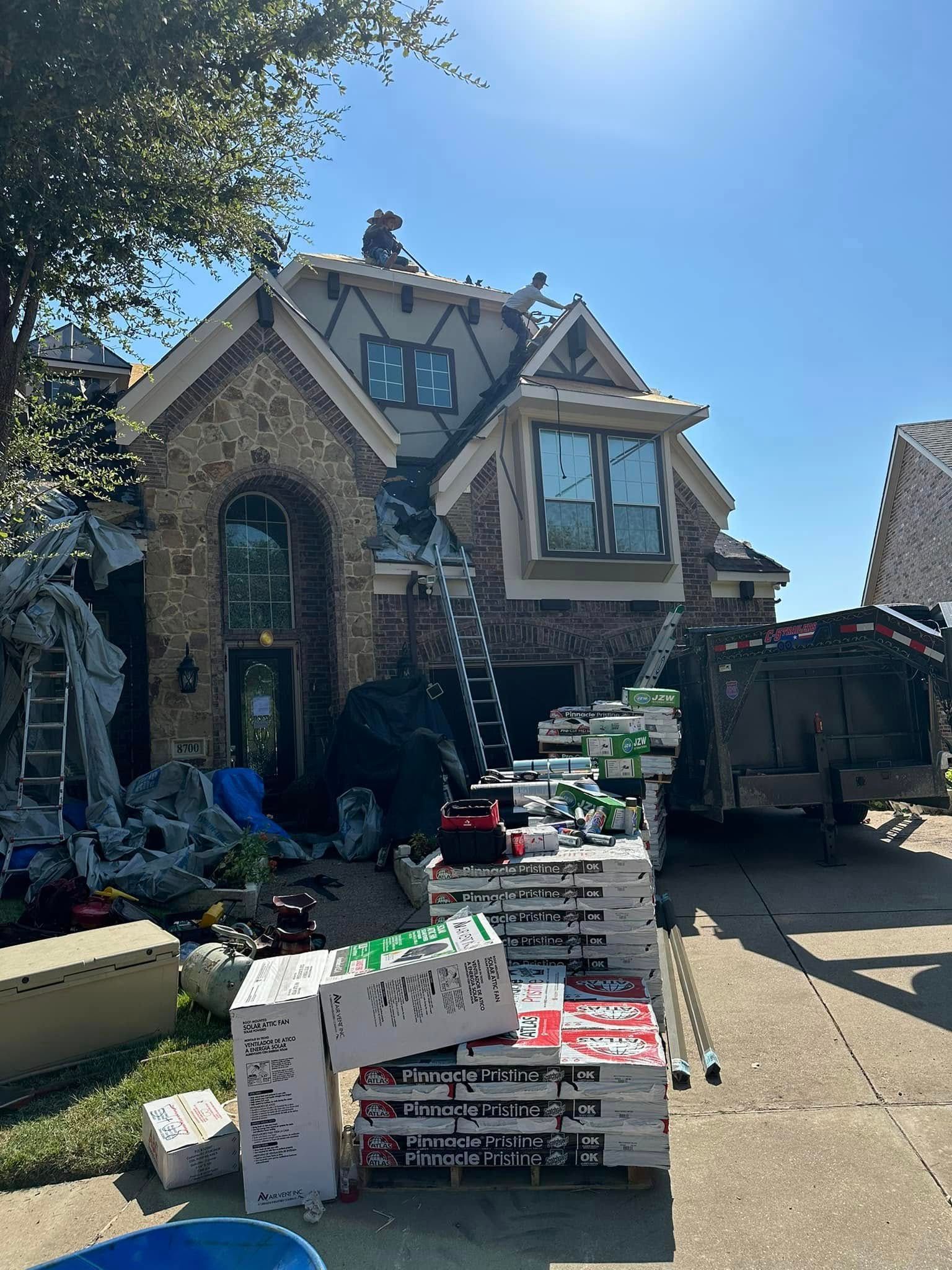 House under construction; roofers working; materials and truck on driveway; blue sky.