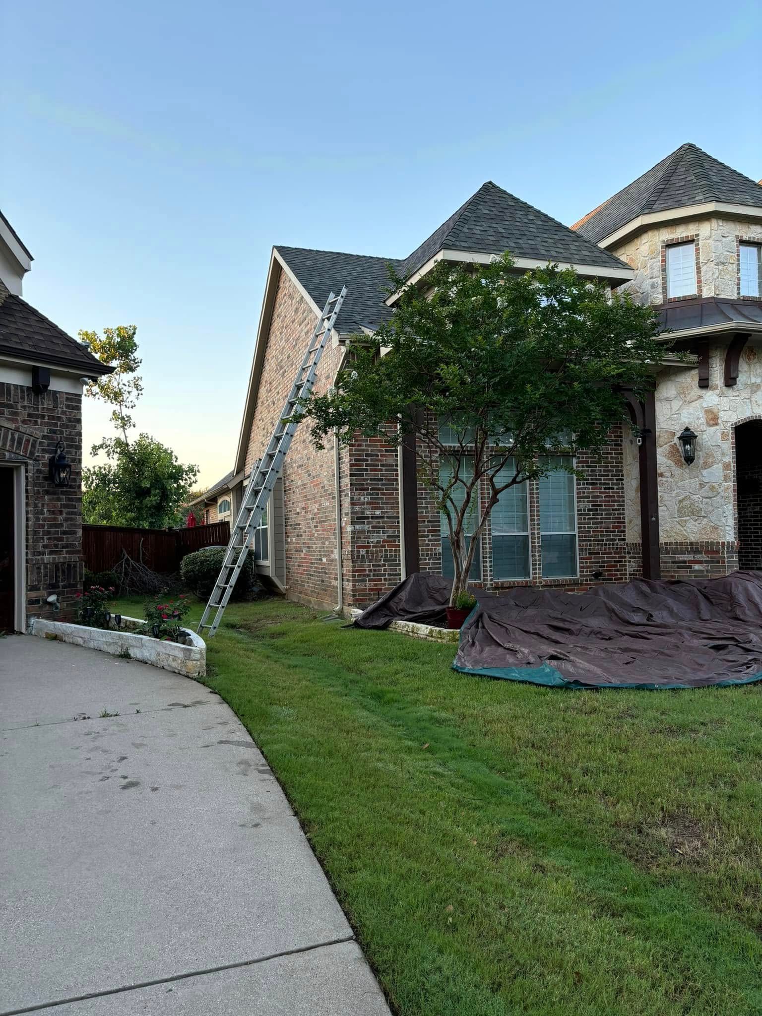 Ladder leaning against a brick house. Green grass, a tarp, and blue sky.