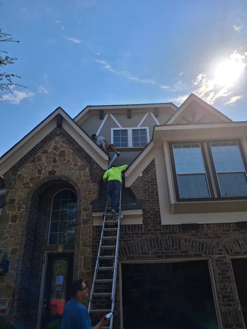 Construction workers on a ladder, working on a house's exterior under a sunny sky.