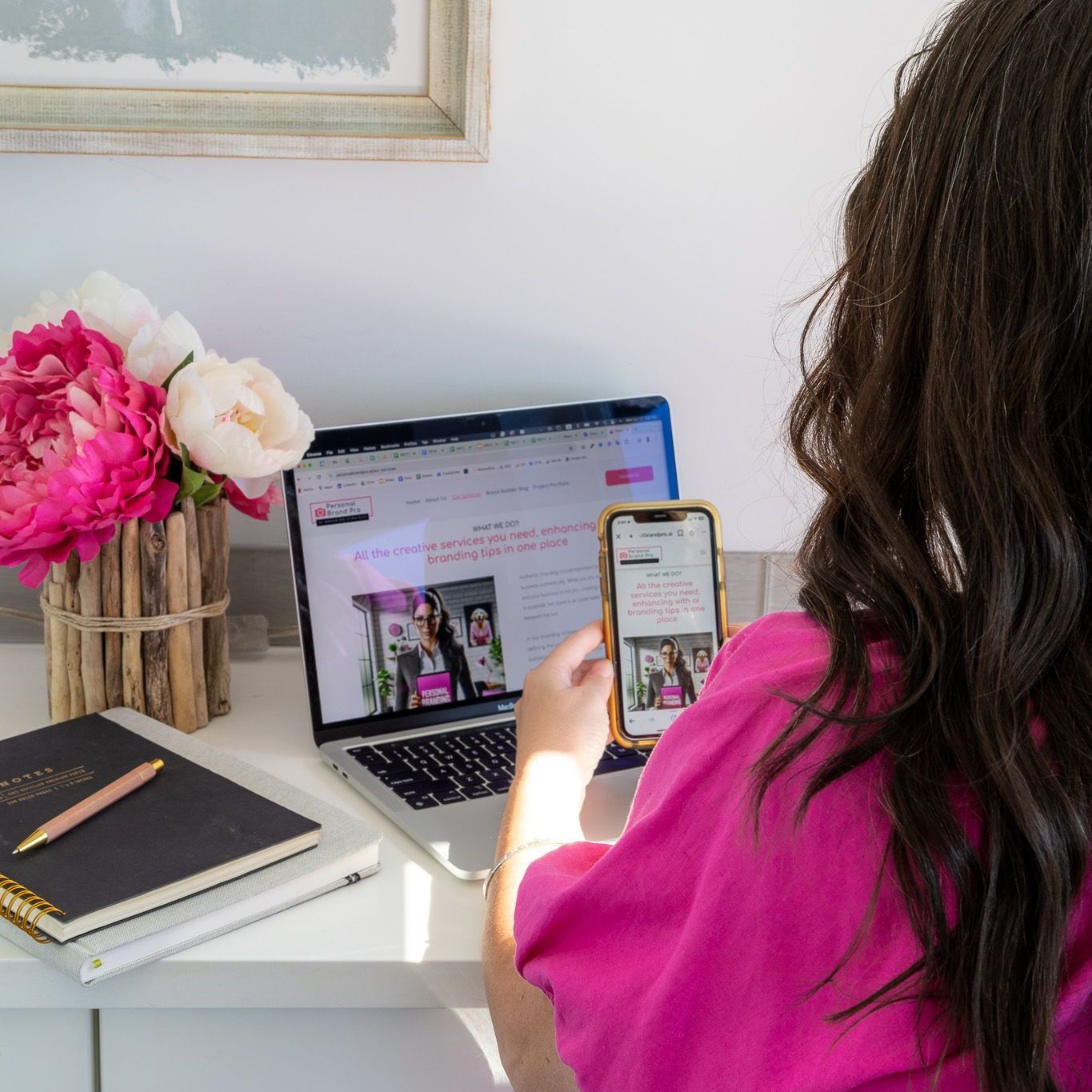 A woman is sitting at a desk using a laptop and a cell phone.