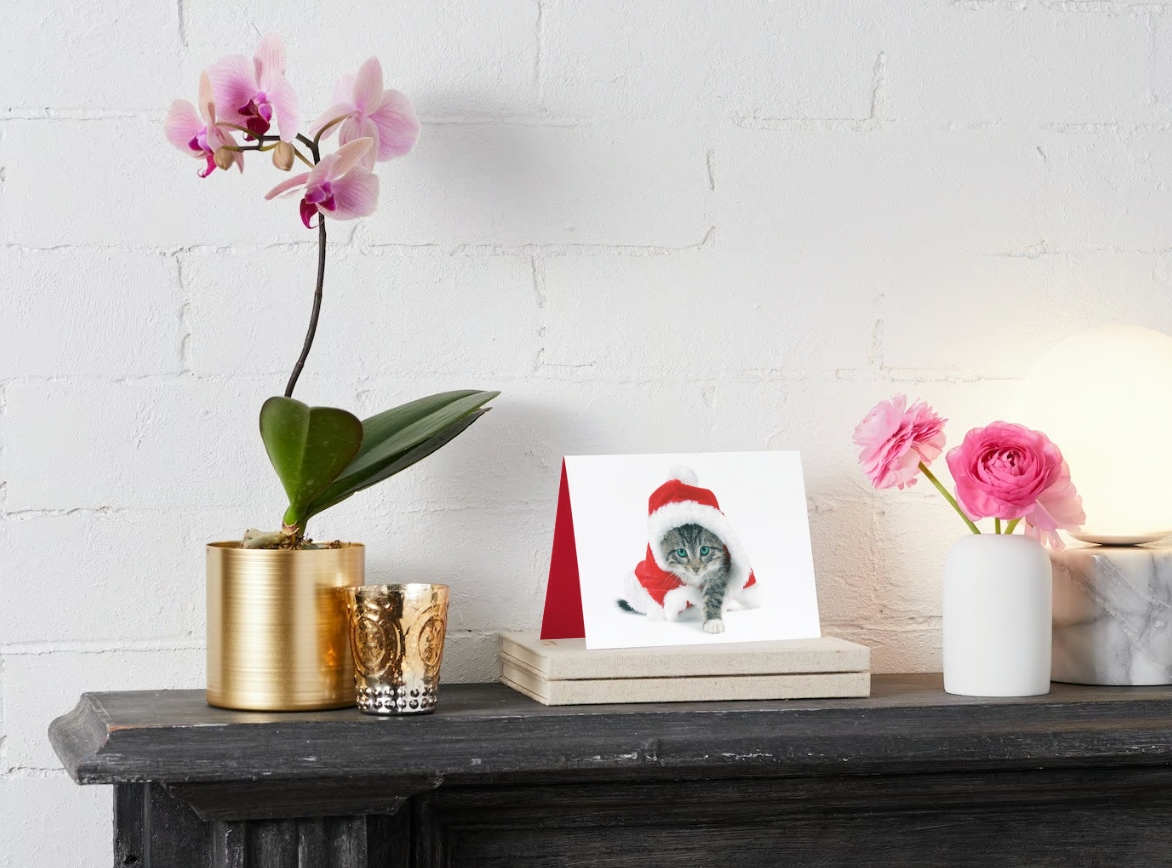 A christmas card with a cat wearing a santa hat is on a shelf next to a vase of flowers.