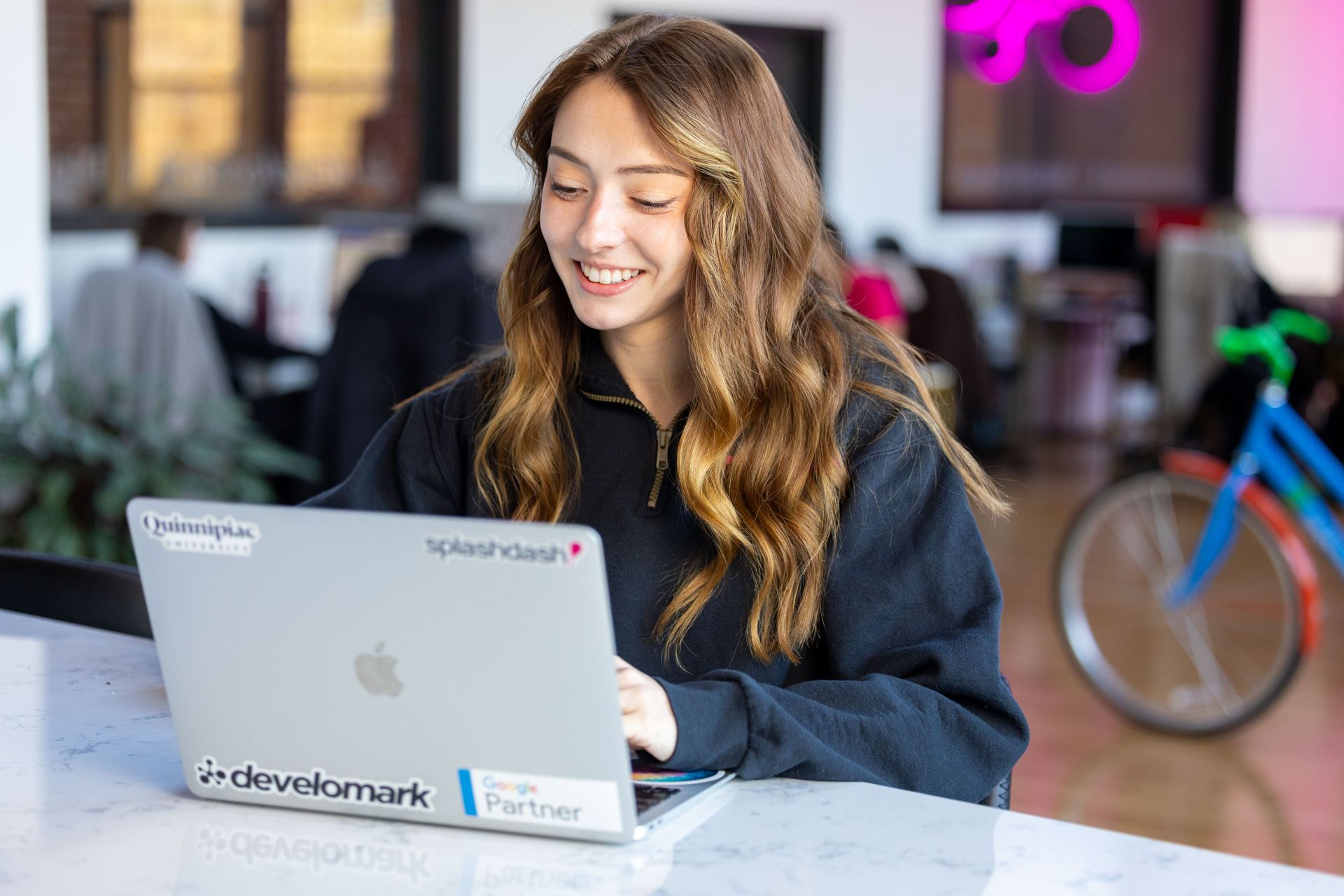Emily Armbruster smiling at a laptop in a bright office, with a bicycle in the background.