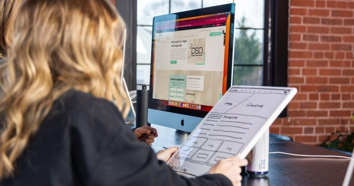 Person using a tablet at a computer workstation, viewing a monitor with a website open.