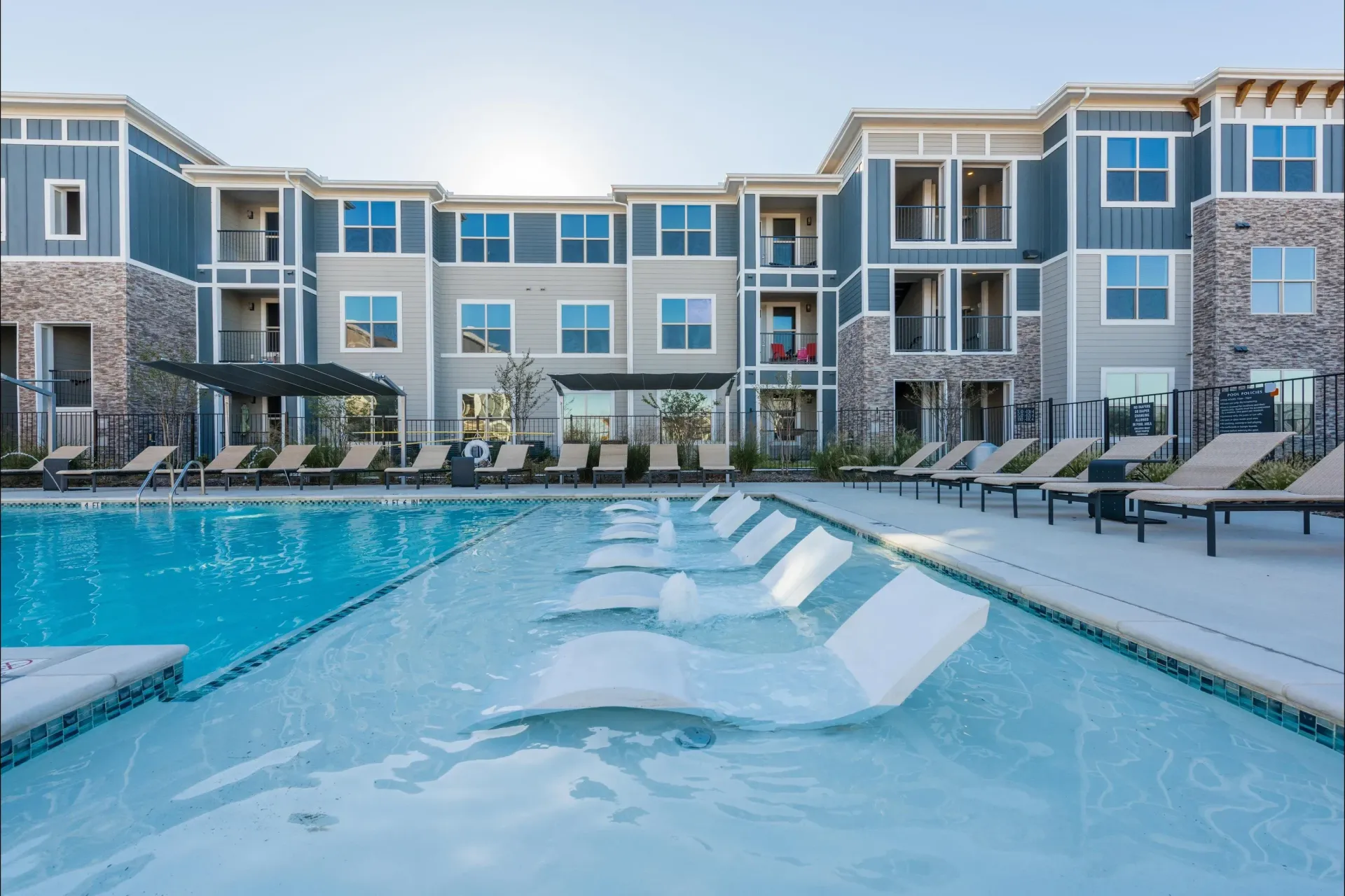 A swimming pool with submerged lounge chairs in front of a modern three-story apartment complex at sunset.
