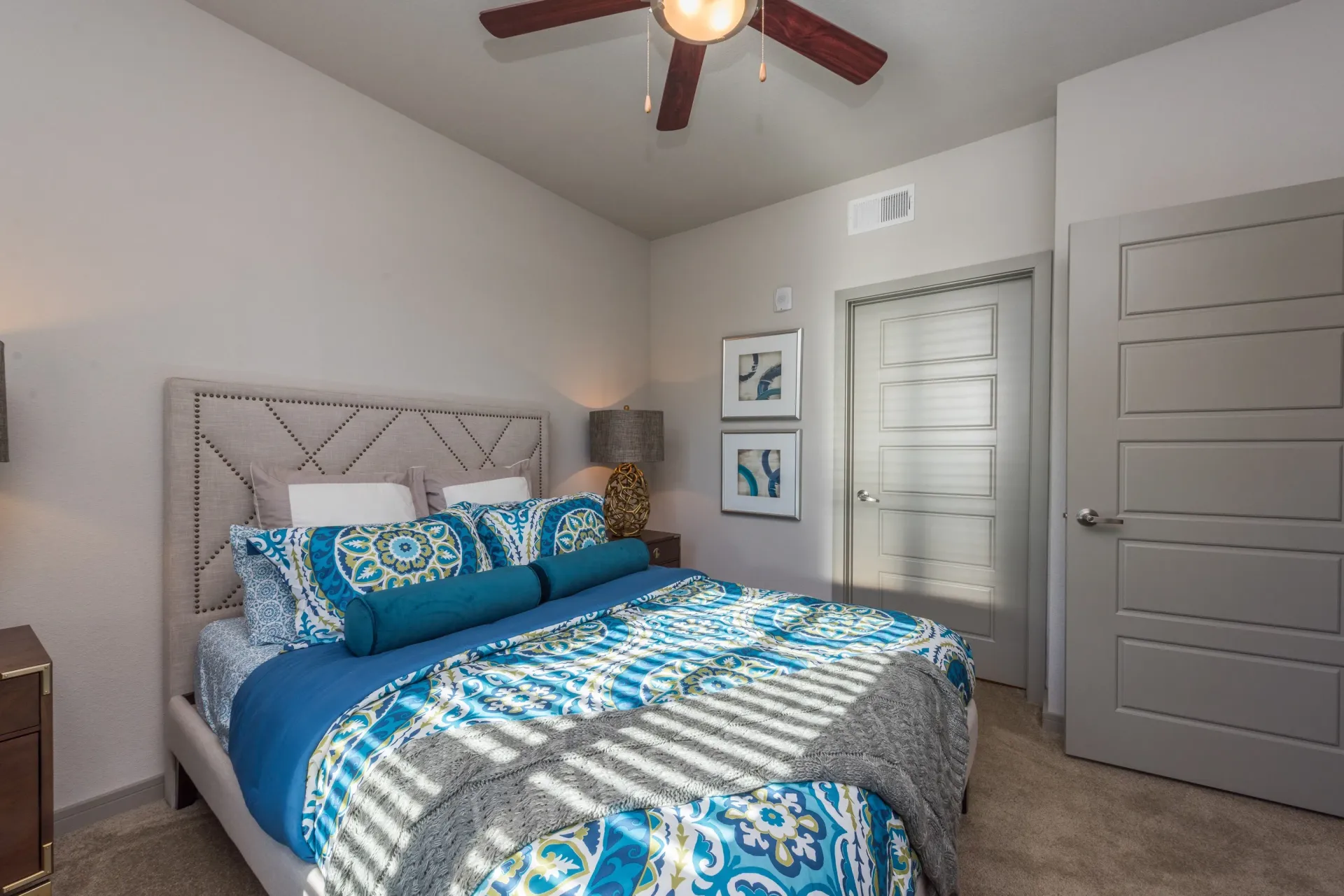 A modern bedroom featuring a tufted beige headboard, blue patterned bedding, wall art, and a gray door.
