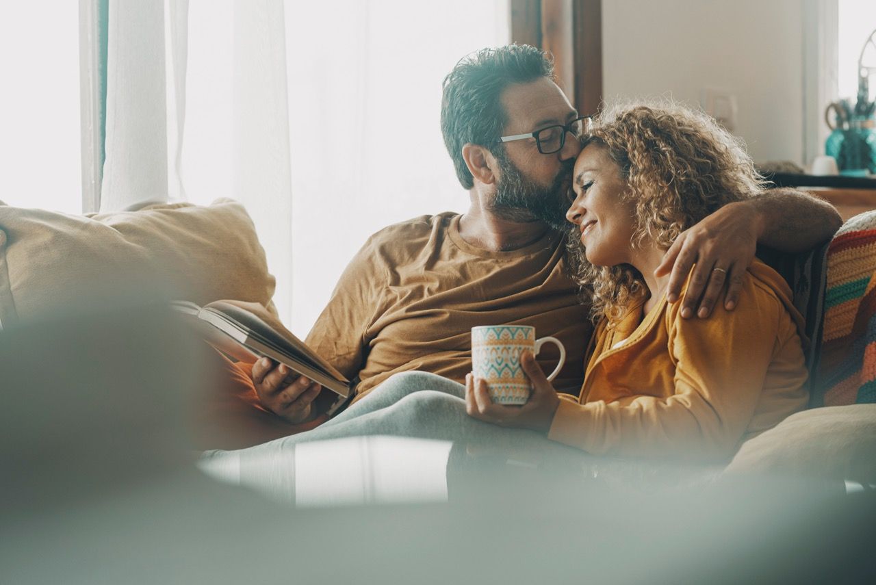 A couple sits together on a couch, one reading a book and holding a mug while the other kisses their head.