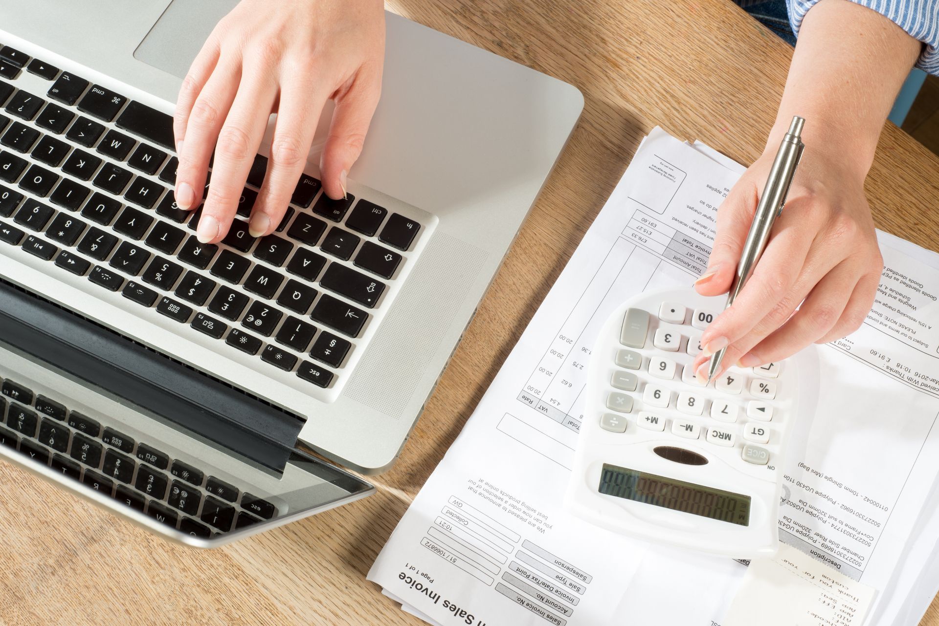 Hands using a calculator and laptop on paperwork spread over a wooden desk.
