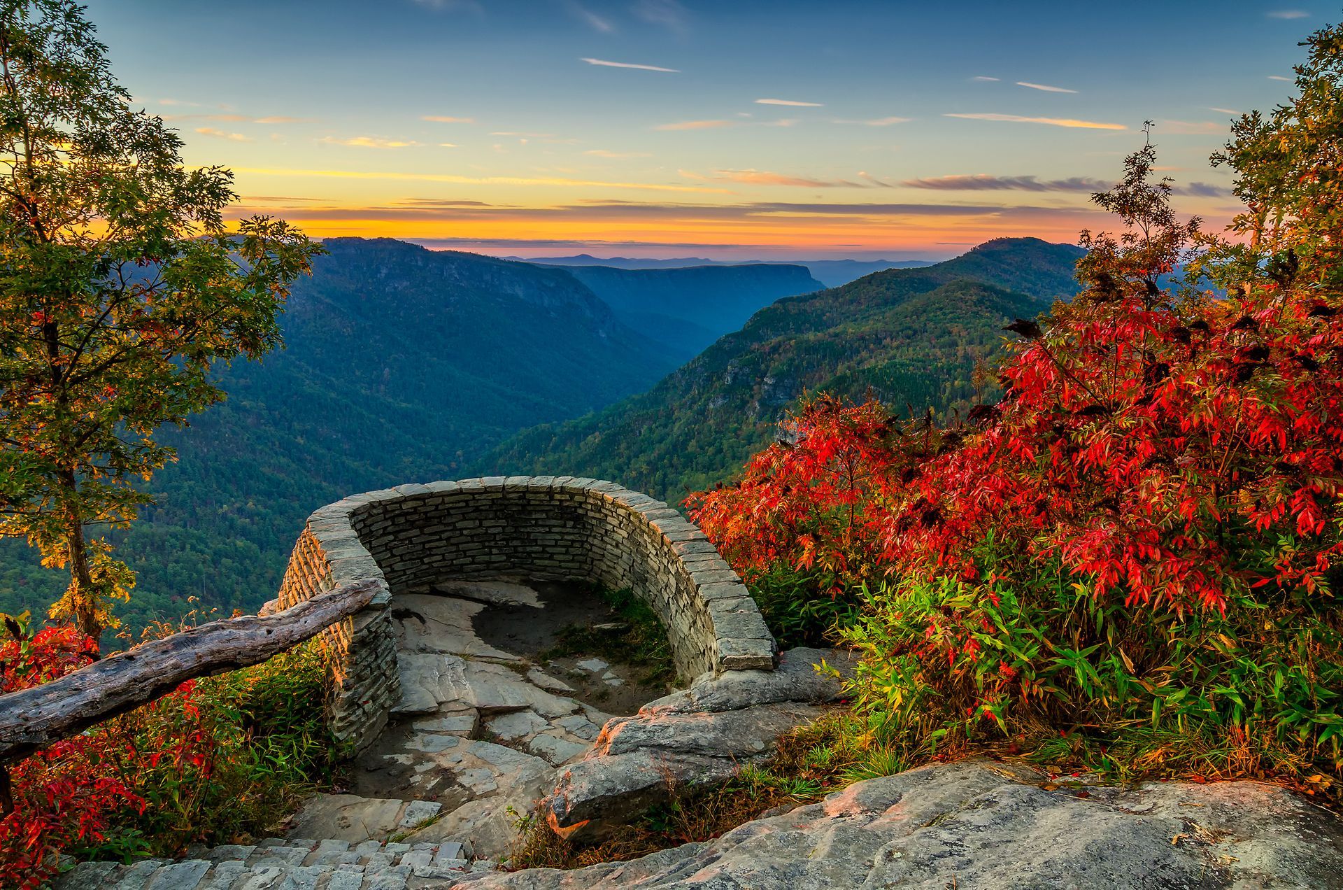 Stone observation deck overlooking a deep, forested valley at sunset with vibrant orange and red autumn foliage.