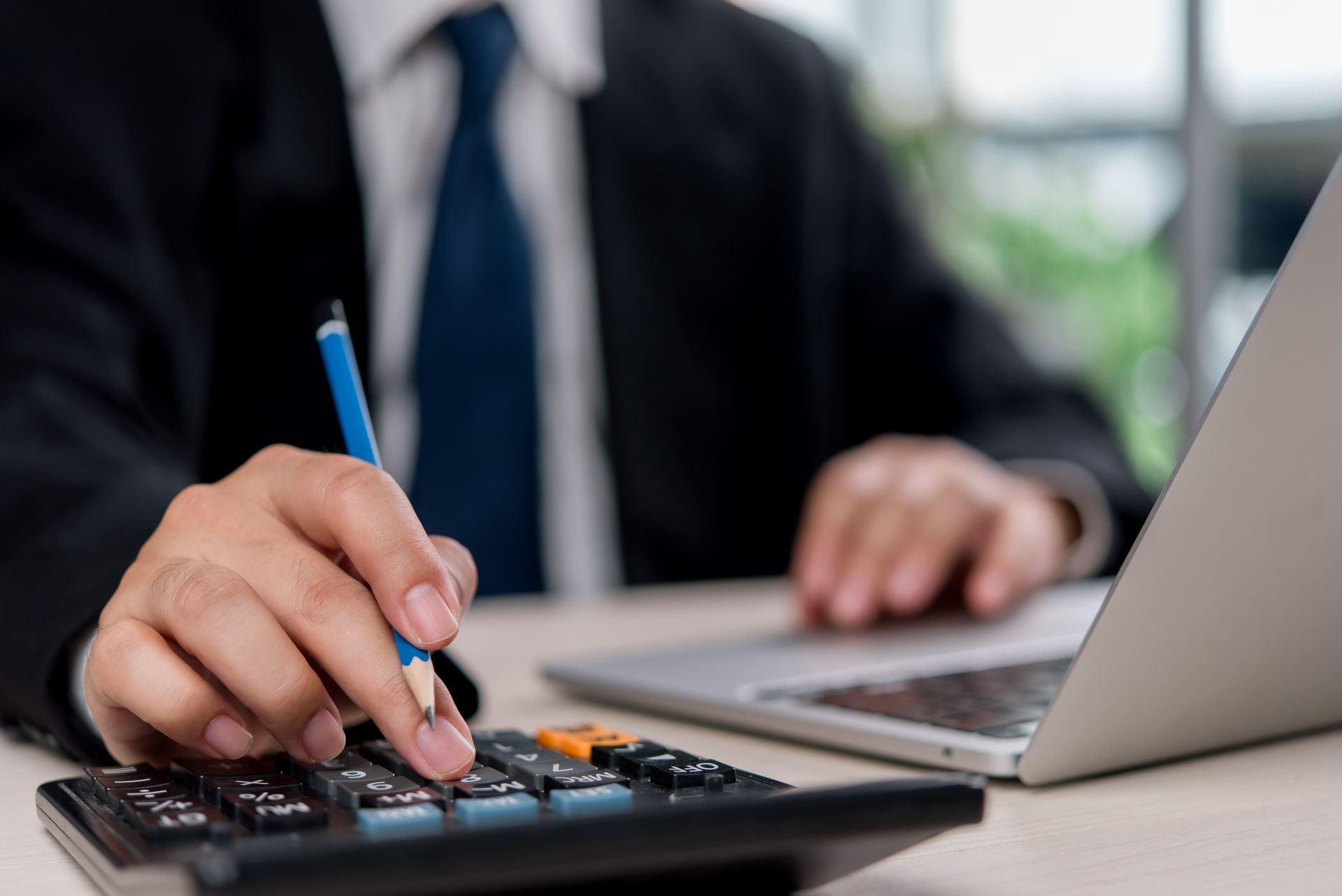 A person in a suit using a calculator and laptop at a desk to perform financial work.