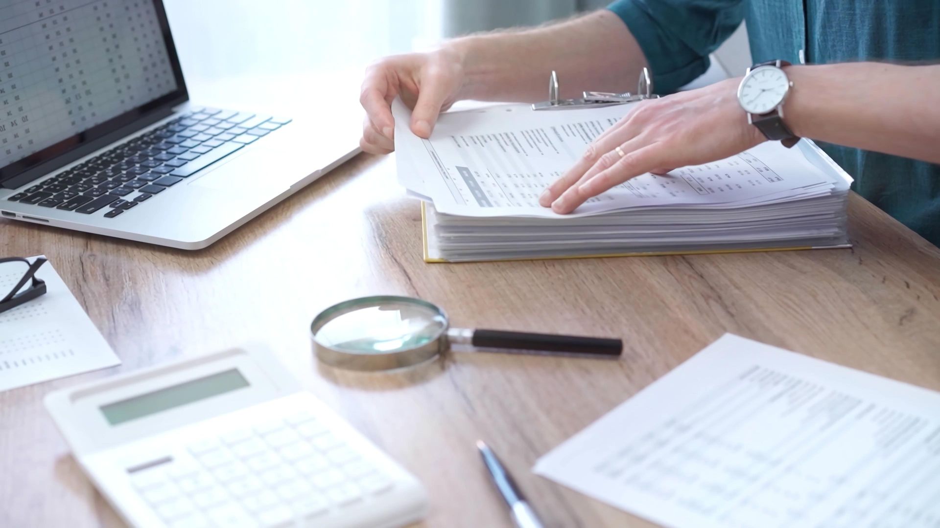 Person working at a wooden desk with a laptop, calculator, magnifier, and a thick binder of documents.
