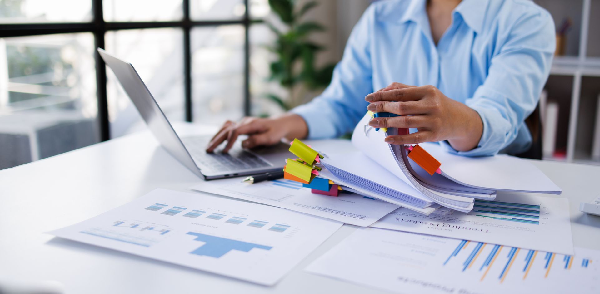 A person in a blue shirt works at a desk with a laptop and documents featuring charts and colorful binder clips.