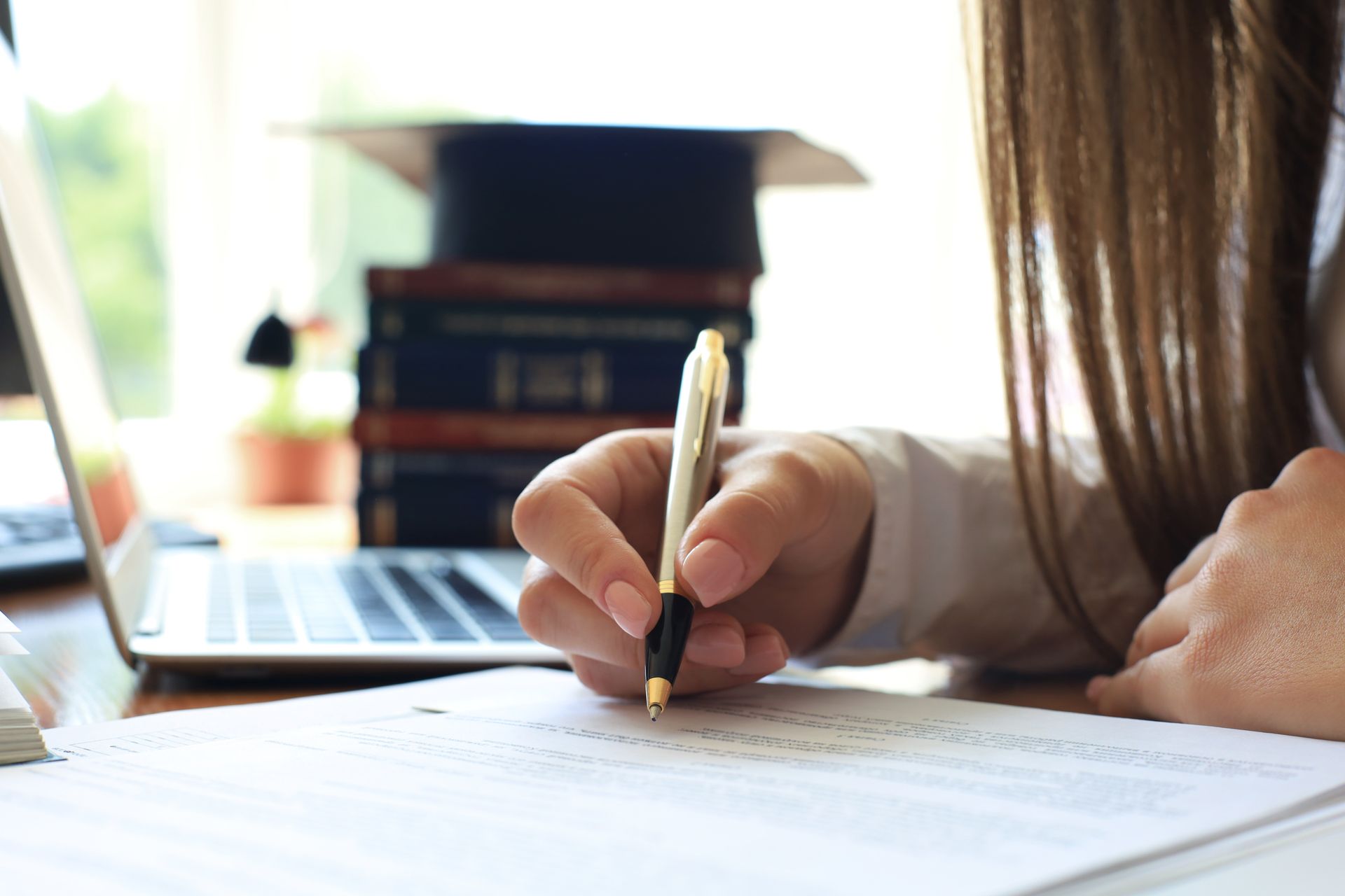 A person holding a pen over a document at a desk with a laptop and a graduation cap on a stack of books in the background.