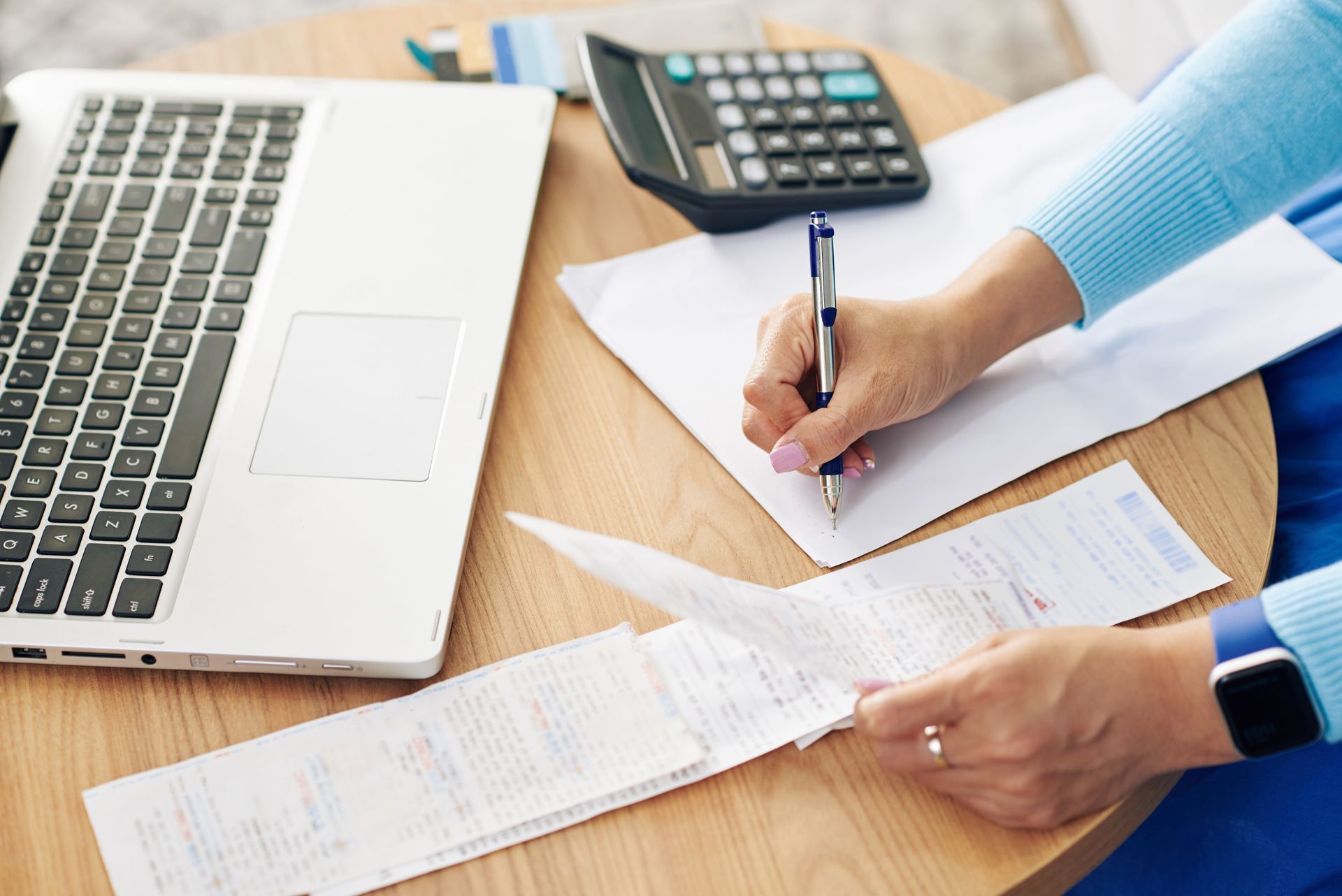 A person sitting at a wooden desk with a laptop and calculator, writing on paper while reviewing a long receipt.