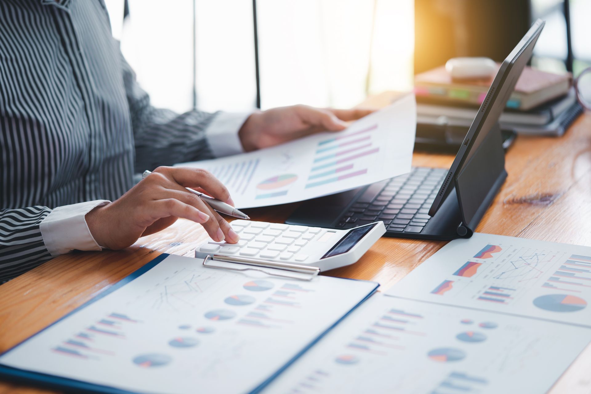 A person at a wooden desk uses a calculator to analyze financial charts, graphs, and reports next to a tablet.