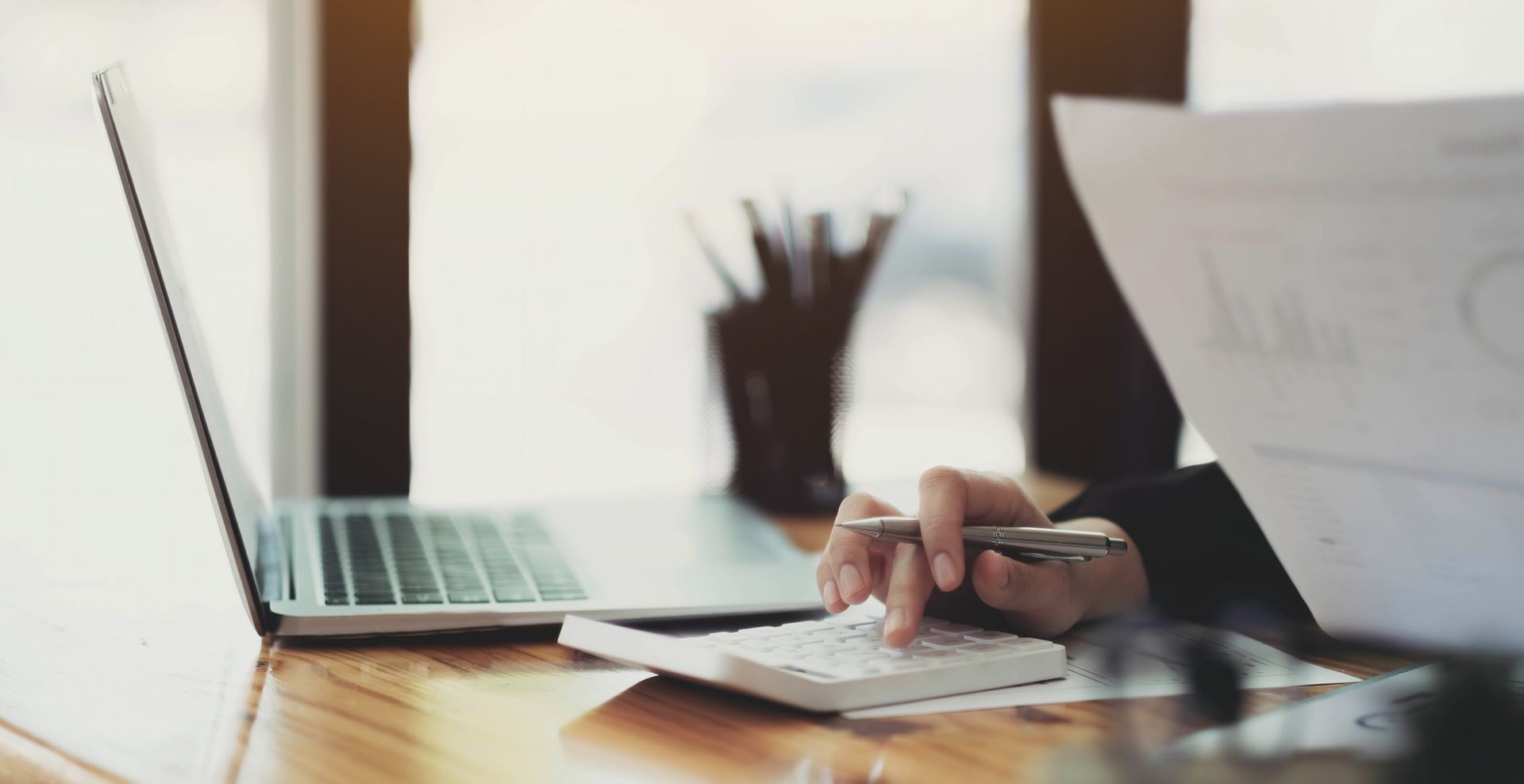 A person works at a wooden desk with a laptop and calculator while reviewing documents in a bright, modern office.