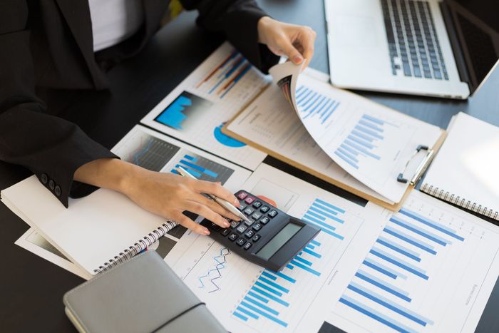 A professional at a desk uses a calculator to analyze financial charts and graphs on printed reports.