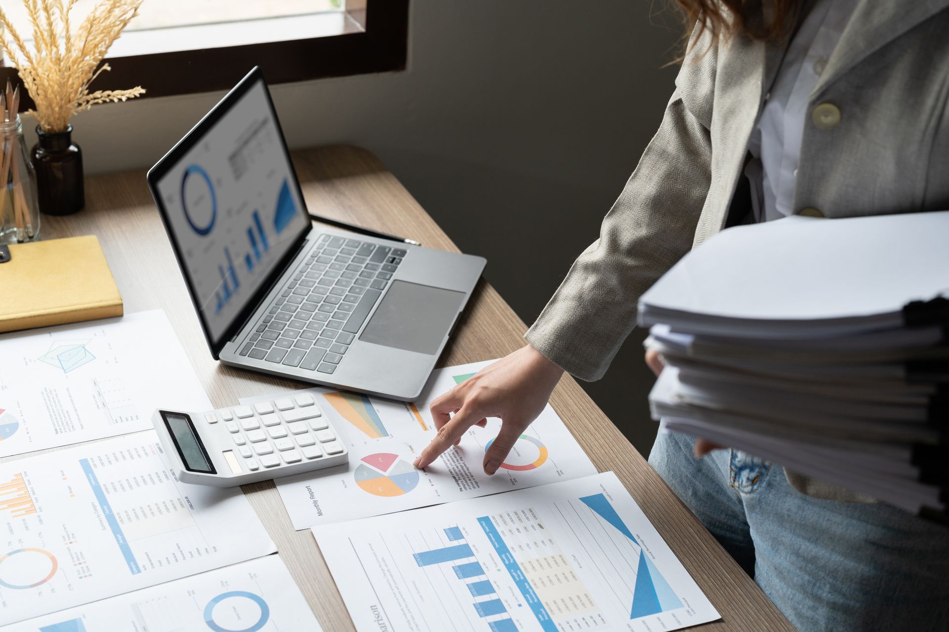 A person standing at a desk with a laptop and documents, pointing to a chart while holding a large stack of papers.