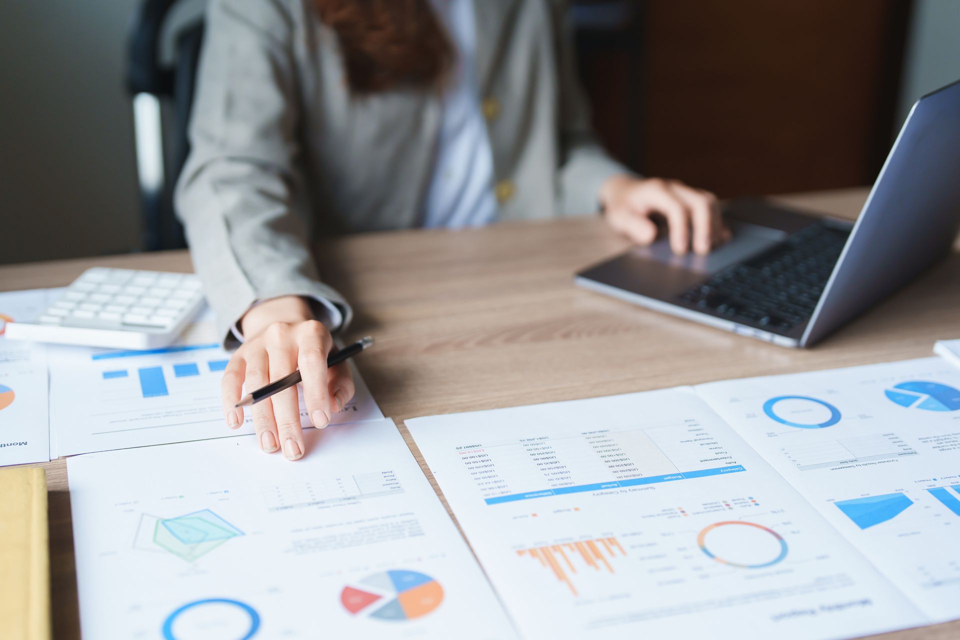 A person in a blazer sits at a desk with financial charts, a laptop, and a calculator, reviewing business data.